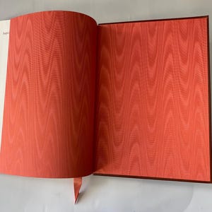 Puede incluir: Un libro abierto con un dise&ntilde;o de patr&oacute;n rojo-naranja en la p&aacute;gina derecha y un margen blanco a la izquierda. El libro tiene una cubierta roja oscura y un marcador de cinta roja.