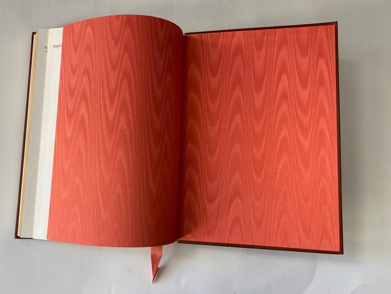 Puede incluir: Un libro abierto con un dise&ntilde;o de patr&oacute;n rojo-naranja en la p&aacute;gina derecha y un margen blanco a la izquierda. El libro tiene una cubierta roja oscura y un marcador de cinta roja.