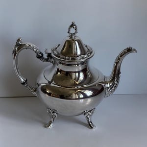 May include: A polished silver teapot featuring a rounded body, a curved spout, and an ornate handle. The lid is topped with a decorative finial, and the base is supported by three scrolled feet. The surface reflects light with a high-gloss finish.
