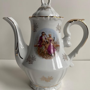 May include: White porcelain teapot with a gold trim and a floral design. The teapot features a painted scene of a couple in 18th-century clothing.