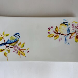 May include: A rectangular white ceramic serving tray featuring two blue and yellow birds perched on flowering branches with green and yellow leaves. The design is painted on the surface with delicate floral details in pink and brown tones.