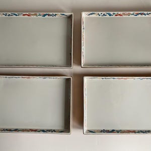 May include: Four rectangular serving platters with a light gray surface and a decorative border. The border features a floral and leaf design in red, blue, and green. The platters are arranged in a square formation.