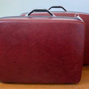 May include: Two vintage red suitcases with silver-toned handles and zippers. The suitcases are rectangular with rounded corners and a textured surface. One suitcase is in front of the other.