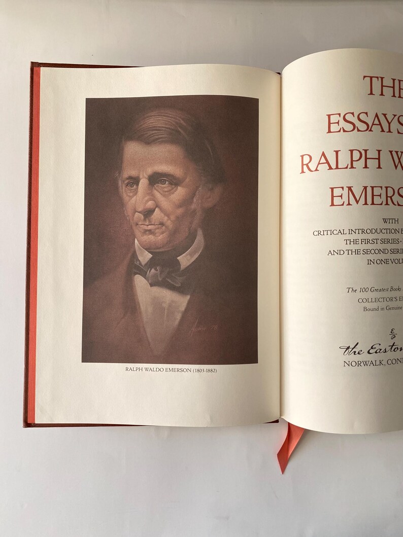 Puede incluir: Un libro abierto con un retrato de Ralph Waldo Emerson en la p&aacute;gina izquierda. La p&aacute;gina derecha presenta texto rojo que dice "The Essays of Ralph Waldo Emerson". Una cinta roja como marcador de libro es visible. El libro est&aacute; encuadernado en una cubierta roja oscura.