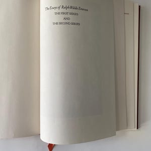 Puede incluir: Libro abierto con el t&iacute;tulo "The Essays of Ralph Waldo Emerson" impreso en la p&aacute;gina derecha. El texto dice "THE FIRST SERIES AND THE SECOND SERIES." Una cinta roja de marcador es visible.