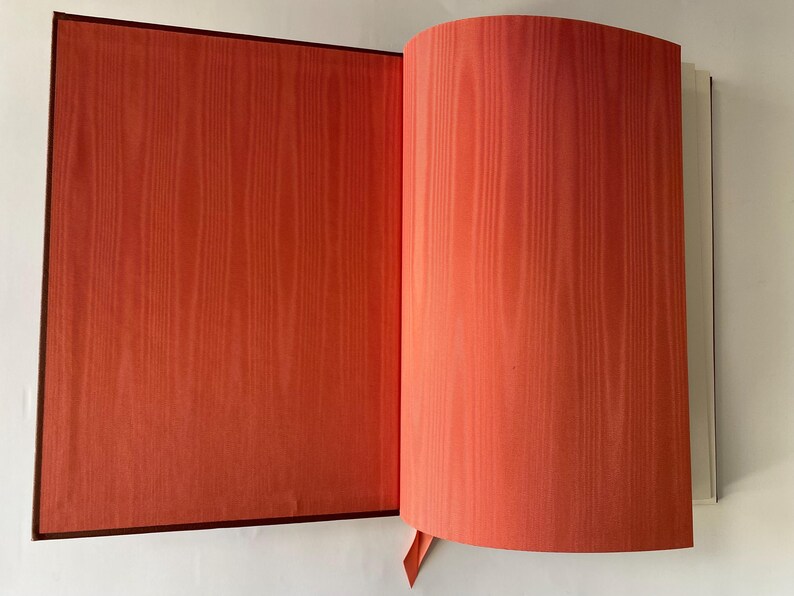 Puede incluir: Un libro abierto con p&aacute;ginas que muestran un patr&oacute;n de vetas de madera de color rojo anaranjado. La cubierta y los bordes del libro son de color marr&oacute;n oscuro. Se ve un marcador de cinta roja.
