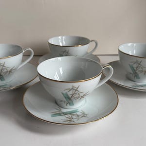 May include: A set of four white porcelain teacups and matching saucers, each featuring a delicate bamboo stalk pattern in shades of green and brown. The rims of both the cups and the saucers are finished with a thin, metallic gold trim.