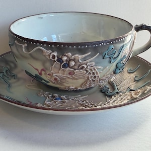 May include: A ceramic teacup and saucer set featuring a raised, three-dimensional dragon motif in shades of brown, blue, and cream. The cup has a curved handle and a smooth white interior, while the exterior and saucer display a detailed dragon.