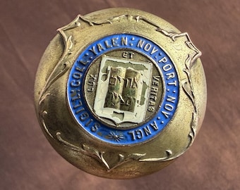 May include: A vintage, round, brass-toned emblem with a blue and white central design. The emblem features text around the perimeter, including "YALE" and "NOV: PORT: NOV". The center design includes a shield with a book and the word "LUX".