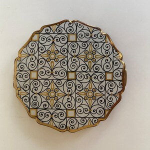 May include: A round, white compact with a gold trim and a black and gold geometric pattern.