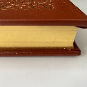 Puede incluir: Primer plano de un libro de tapa dura con una cubierta de cuero marr&oacute;n y un dise&ntilde;o en relieve dorado. Las p&aacute;ginas son de color amarillo dorado y los bordes est&aacute;n dorados. El libro est&aacute; sobre una superficie blanca.