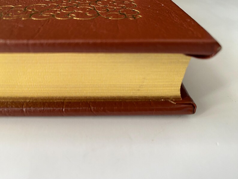 Puede incluir: Primer plano de un libro de tapa dura con una cubierta de cuero marr&oacute;n y un dise&ntilde;o en relieve dorado. Las p&aacute;ginas son de color amarillo dorado y los bordes est&aacute;n dorados. El libro est&aacute; sobre una superficie blanca.