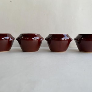 May include: Four maroon-colored ceramic bowls with a glossy finish. Each bowl has a unique shape with a wide opening, a slightly tapered body, and small handles on the sides. The bowls are arranged in a row.