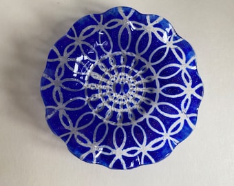 May include: A round, blue glass decorative plate with a scalloped edge. The plate features a silver, geometric floral pattern radiating from the center. The plate is approximately 8 inches in diameter.