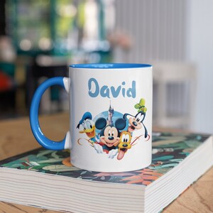 May include: A white ceramic mug with a blue rim and handle. The mug features a colorful illustration of Mickey Mouse, Donald Duck, Goofy, and Pluto. The name "David" is written in blue cursive on the mug.