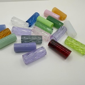 May include: A collection of colorful, cylindrical glass beads in various shades, including blue, green, purple, and amber. Each bead has a textured, ridged surface, and they are scattered on a white surface.