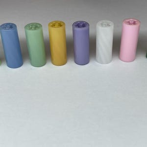May include: Eight small cylindrical tubes in various colors, including green, blue, yellow, purple, white, pink, and dark green. The tubes are arranged in a row on a white surface.