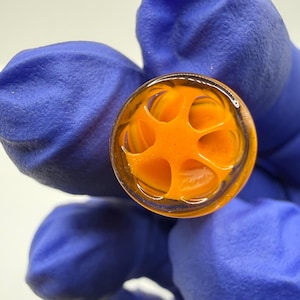 May include: Close-up of a clear glass object with an orange, star-shaped design inside. The object is held by a person wearing blue gloves. The glass has a circular shape and is filled with a vibrant orange color.
