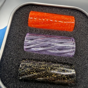 May include: Three cylindrical objects arranged vertically in a foam-lined metal tin. The top cylinder is bright orange with gold flecks, the middle is translucent light purple with a twisted texture, and the bottom is black with gold flecks.