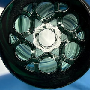 May include: A circular glass object featuring a dark green outer rim and a central white hexagonal core. The interior is filled with a pattern of translucent green and white circular and triangular shapes that create a layered, reflective effect.