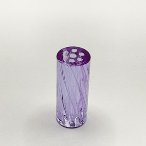 May include: A cylindrical, purple glass bottle stopper with a swirl design. The top of the stopper has six small holes. The glass has a transparent quality, allowing light to pass through. The stopper is designed for use with bottles.