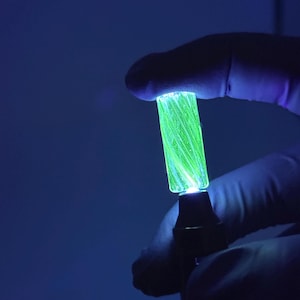 May include: A glowing, cylindrical object held by a gloved hand against a dark blue background. The object emits a bright green light, with visible internal structures. The base is dark, and the overall effect is a striking contrast of light and shadow.