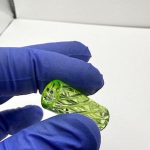 May include: A small, cylindrical, green glass object with a spiral design. The object is held between two hands wearing blue gloves. The glass has a textured surface and a hole at one end.