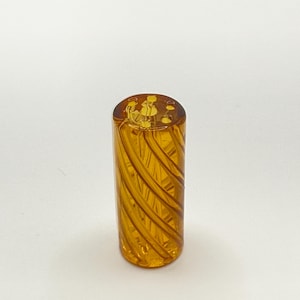 May include: A cylindrical, amber-colored glass object with a spiral design. The glass has a glossy finish and appears to be hand-blown. The object is standing upright against a white background. The top of the object has a decorative design.