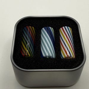 May include: Three glass cylinders with colorful, diagonal stripes are displayed in a small, square metal tin. The cylinders have stripes in various colors, including blue, red, yellow, and white. The tin is lined with black foam.