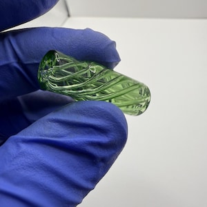 May include: A green glass object with a spiral design. The object is cylindrical with rounded ends and appears to be hand-blown. The glass has a slight tint of green and is held by a person wearing blue gloves.