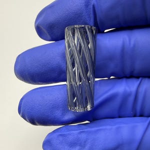 May include: A clear, cylindrical glass object with a spiraling, interwoven design. The object is held by a person wearing blue gloves. The glass has a textured appearance, with light reflecting off the angled surfaces.