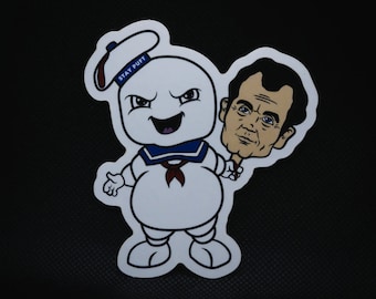 May include: A white cartoon ghost with a blue sailor hat and red scarf holding a man's head. The ghost has the words "Stay Puft" on its hat.