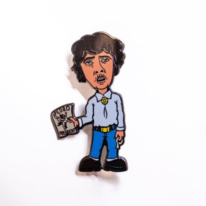 Może przedstawiać: Emaliowana przypinka z postacią kreskówkowego mężczyzny o brązowych włosach, ubranym w jasnoniebieską koszulę, niebieskie dżinsy i żółty pasek. Trzyma w ręku białą ulotkę z napisem "Pedro Prezydent".