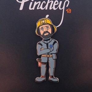 May include: Enamel pin featuring a cartoon character in a gray jumpsuit and yellow helmet. The character has a beard and crossed arms. The pin has a black background with the word "Pinchey" in white script.