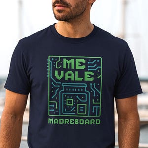 May include: Navy blue t-shirt with a green and teal graphic. The graphic features the words "ME VALE" above a circuit board design and the word "MADREBOARD" below. The design has a pixelated aesthetic.