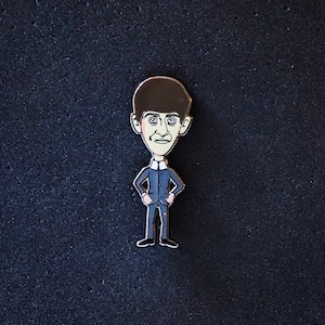 May include: An enamel pin featuring a cartoon man with a large head and a dark blue suit. The man has short black hair and a light brown complexion.
