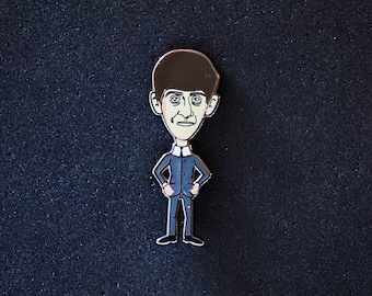 May include: A cartoon enamel pin of a man with a large head and a dark blue suit. The man has short black hair and a light brown complexion.