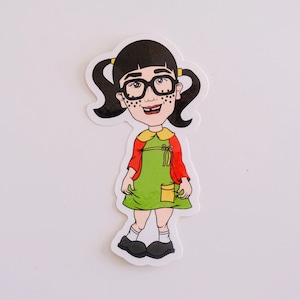 May include: A sticker of a cartoon girl with black hair, wearing glasses, a red and green dress, and black shoes. She has black dots on her face.