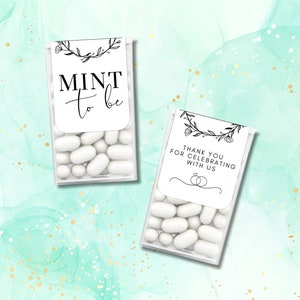 May include: Two small clear plastic containers filled with white mints. The containers have white labels with black text and floral designs. The label on the left container reads "Mint to be". The label on the right container reads "Thank you for celebrating with us" with a small illustration of two rings.