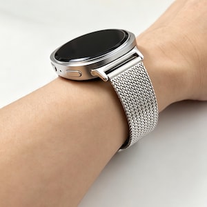 May include: A silver-toned smartwatch featuring a round, black digital display and a metallic mesh strap. The watch case has a smooth, circular bezel with a side button and a small microphone port visible on the side of the metallic housing.