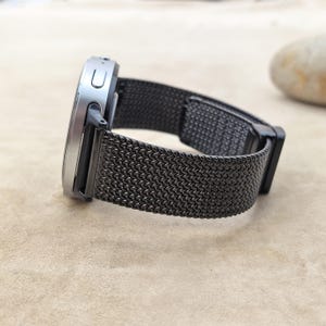 May include: A smartwatch with a metallic silver casing and a dark charcoal gray Milanese loop mesh band. The side of the watch features a rectangular button and a small circular port. The band has a fine, woven texture and a secure clasp mechanism.