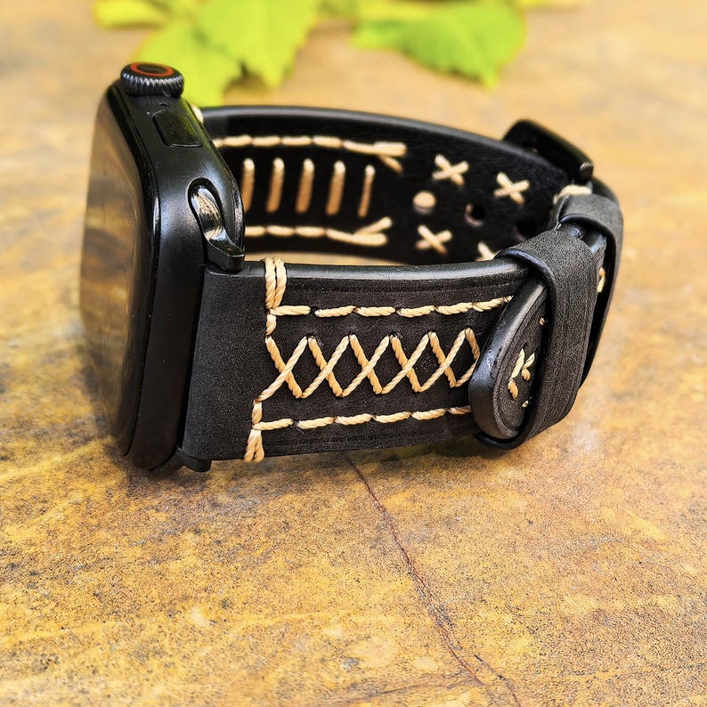Op de afbeelding: Zwart leren Apple Watch band met witte stiksels. Het band heeft een decoratief stikselpatroon en een gespsluiting.