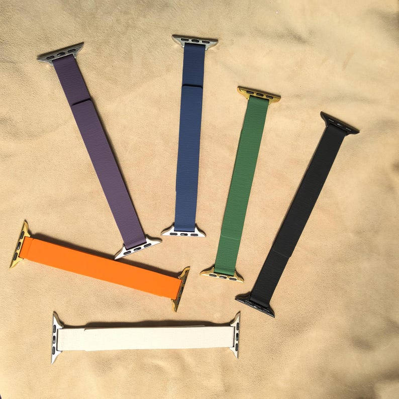 May include: A collection of Apple Watch bands in various colors, including purple, blue, green, black, orange, and white. The bands have metal connectors in silver and gold tones.