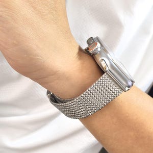 May include: A silver metal watch band with a woven mesh design. The band is worn on a person's wrist.