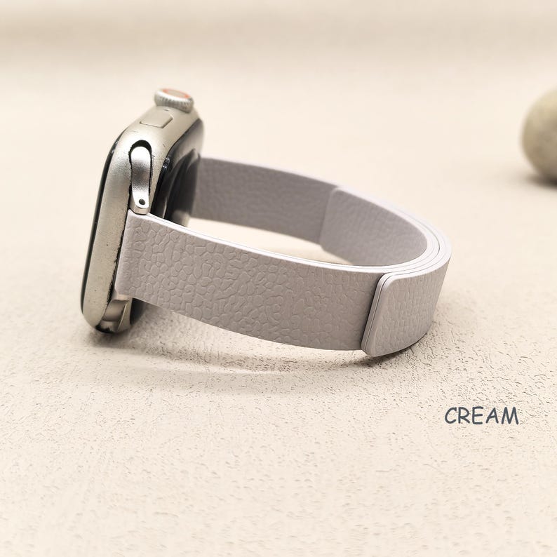 May include: A cream-colored smartwatch with a light gray leather band. The band has a textured surface and is connected to the silver watch face. The word "CREAM" is printed on the surface.