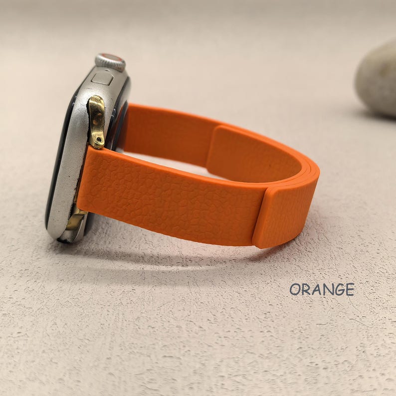 May include: An orange watch band is attached to a silver watch face. The band is made of a textured material and is curved. The word "ORANGE" is printed in white on the surface.