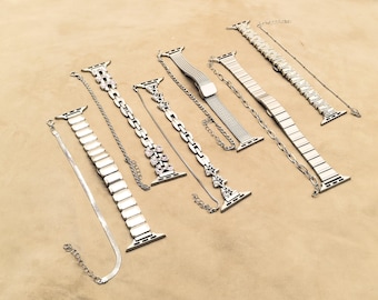 May include: An assortment of silver-toned watch bands, including chain, mesh, and link styles. Some bands feature decorative embellishments like crystals. The bands are displayed on a neutral, tan-colored surface.