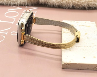 Puede incluir: Una correa de reloj de malla de acero inoxidable dorada sujeta a un reloj inteligente plateado y dorado. La correa está conectada al reloj con herrajes dorados. El reloj está sobre una superficie clara y texturizada.