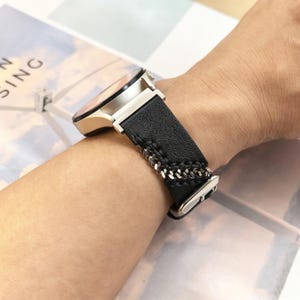 Op de afbeelding: Een smartwatch met een ronde metalen behuizing en een zwarte, gestructureerde leren band. De band is voorzien van decoratieve zilverkleurige kettingdetails die in het leer zijn verwerkt met zichtbare stiksels langs de randen.