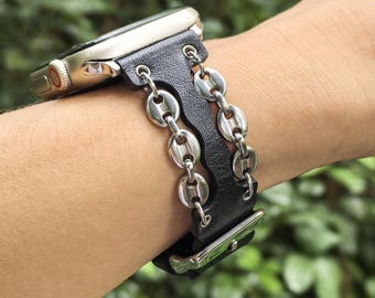 May include: A black leather Apple Watch band with silver-colored chain links. The watch has a silver case and is attached to the band. The band is worn on a wrist, with a blurred green background.
